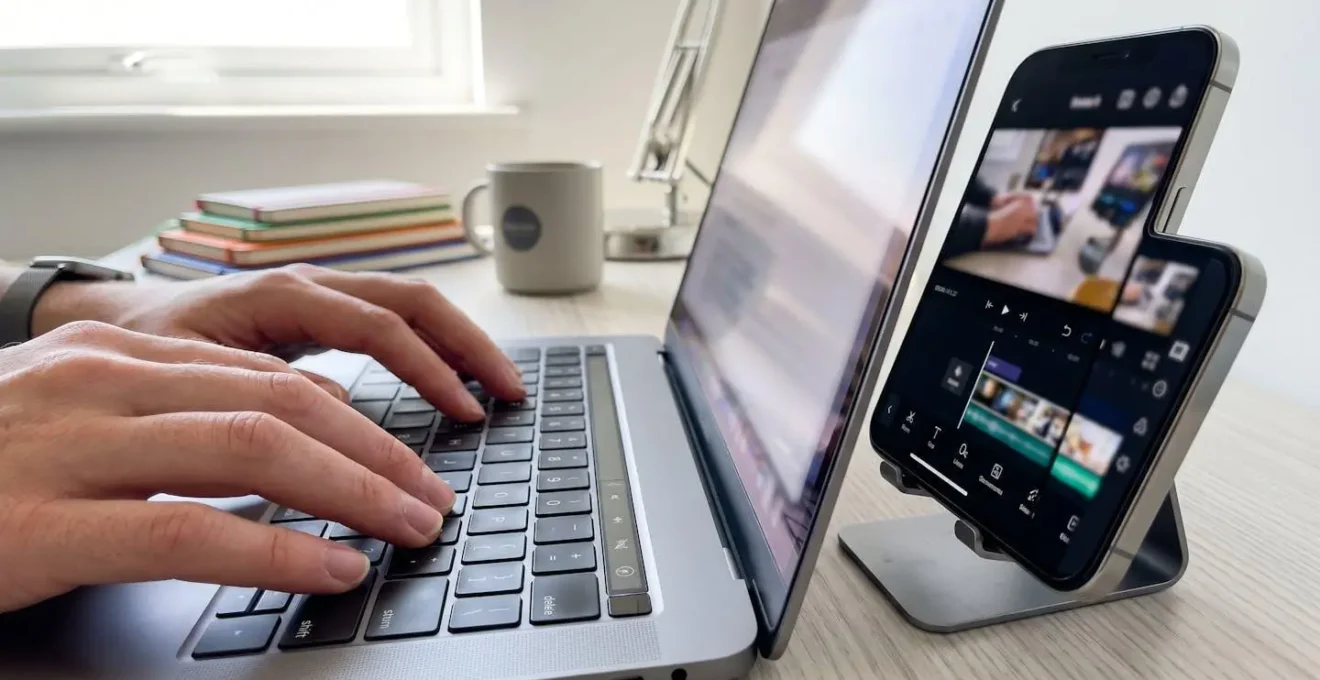 Close-up of hands typing on keyboard with smartphone displaying video creation interface beside laptop on modern desk